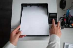 Hand holding magnifying glass over A4 paper in printer tray to diagnose faults like banding/streaking: Poor Print Quality Diagnostics.