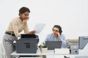An exasperated employee is bent over a photocopier, trying to clear a paper jam