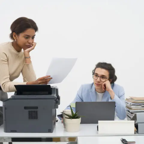 An exasperated employee is bent over a photocopier, trying to clear a paper jam