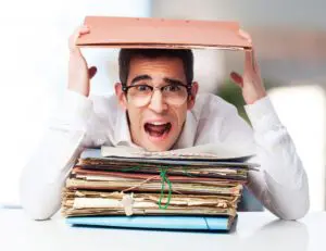 A frustrated office worker sits at a desk overwhelmed by massive piles of paperwork and disorganized documents.