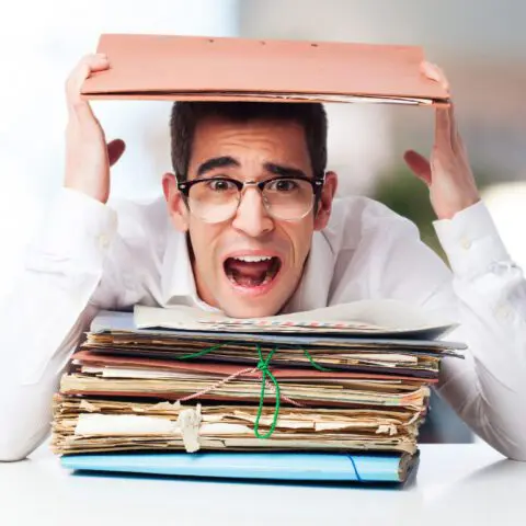 A frustrated office worker sits at a desk overwhelmed by massive piles of paperwork and disorganized documents.