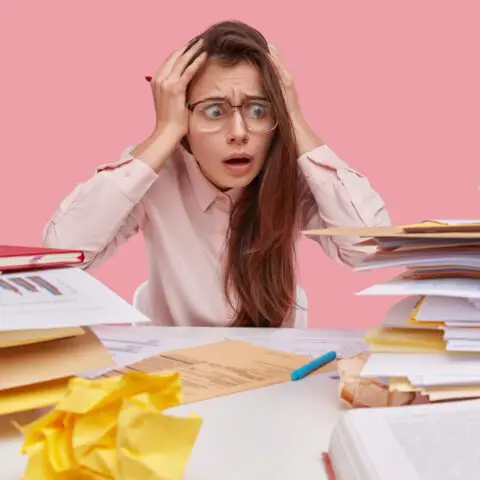 Stressed office worker at a desk overwhelmed by towering piles of paperwork and documents.