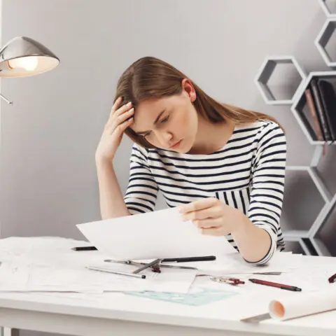 An office worker looks stressed while sitting at a desk surrounded by large piles of paperwork and files.