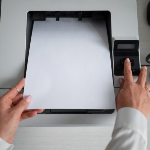 Hand holding magnifying glass over A4 paper in printer tray to diagnose faults like banding/streaking: Poor Print Quality Diagnostics.