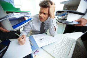 Frustrated office worker sitting at a desk covered in stacks of paper, struggling to locate a file.