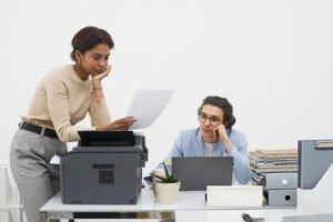 An exasperated employee is bent over a photocopier, trying to clear a paper jam