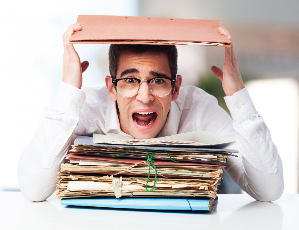 A frustrated office worker sits at a desk overwhelmed by massive piles of paperwork and disorganized documents.
