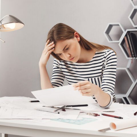 An office worker looks stressed while sitting at a desk surrounded by large piles of paperwork and files.