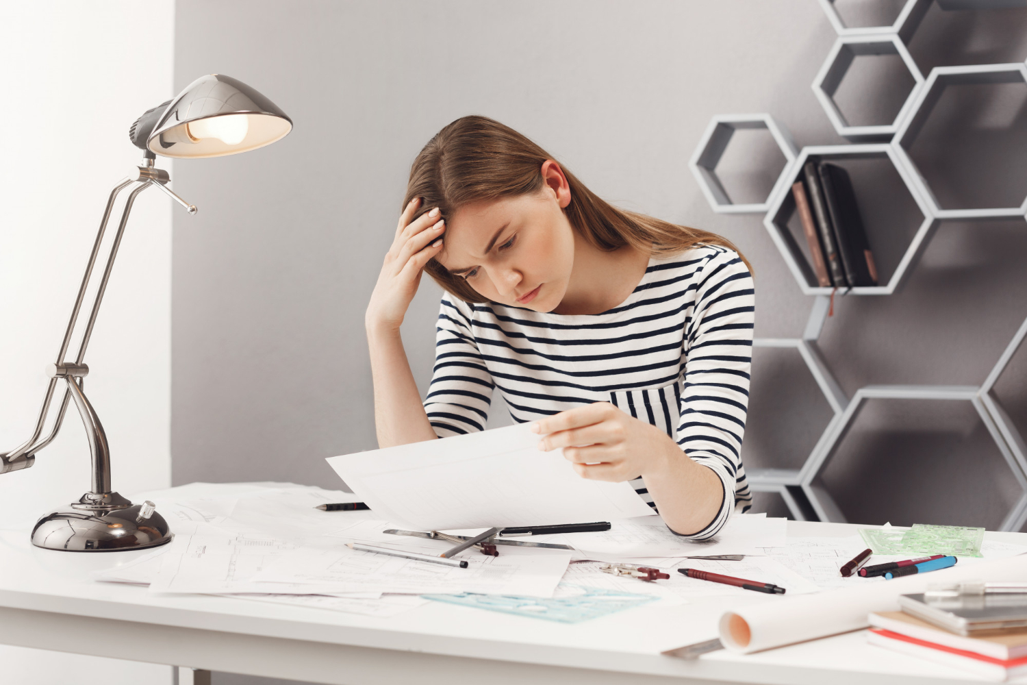 An office worker looks stressed while sitting at a desk surrounded by large piles of paperwork and files.