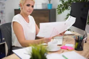 An office worker looks stressed while sitting at a desk surrounded by large piles of paperwork and files.