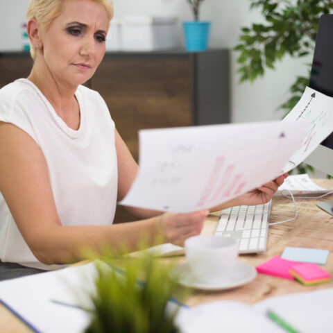 An office worker looks stressed while sitting at a desk surrounded by large piles of paperwork and files.