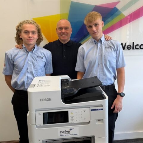 Two young technical apprentices work together inspecting and repairing modern office printer equipment.